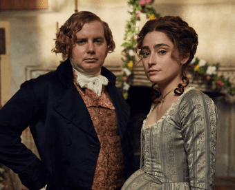 Ellise Chappell with her fellow actor from Poldark in 19th-century attire standing side-by-side and looking directly at the camera.