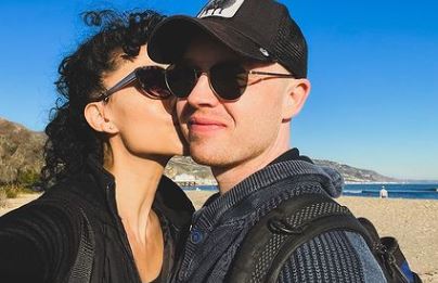 A selfie Noel Fisher with his wife Layla where Layla kisses him on the cheeks and him smiling looking at the camera.