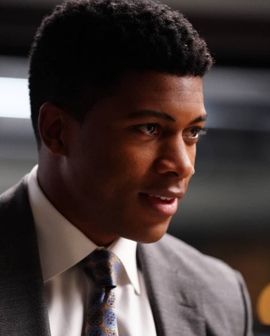A close-up profile shot shows Eli Goree smiling slightly while wearing a grey suit jacket, white shirt, and a patterned blue and gold tie.