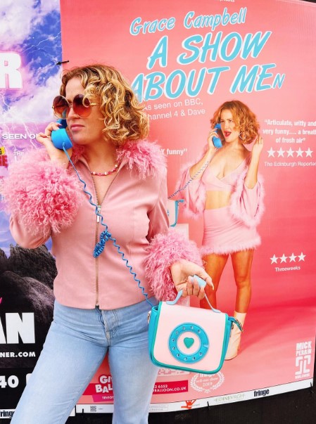 Grace Campbell promoting her show on social media where she holds a retro blue phone to her ear while standing in front of a poster for "Grace Campbell, A SHOW ABOUT MEN."