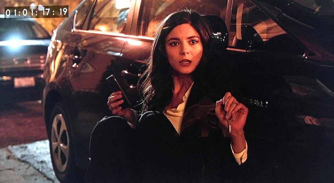 A shot from the show Chicago Justice shows actress Monica Barbaro, sitting on the ground next to a car, holding her phone and looking up with a surprised expression.