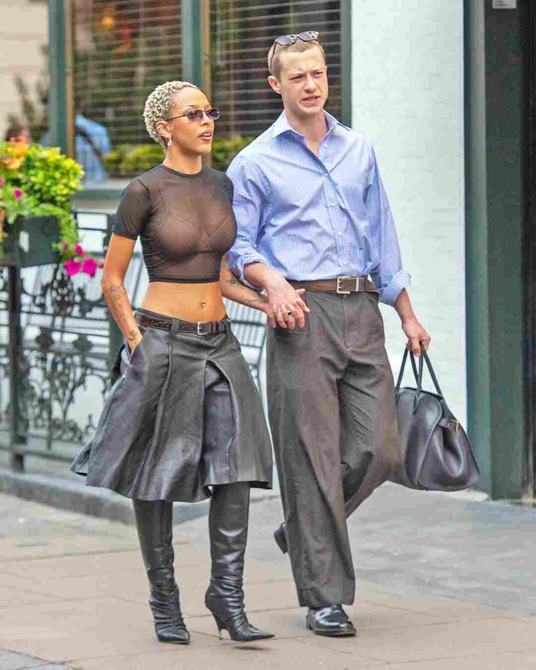 A photo of Joseph Quinn and Doja Cat walking together outdoors, with Joseph wearing a blue shirt and gray pants and Doja Cat wearing a black top and leather skirt.