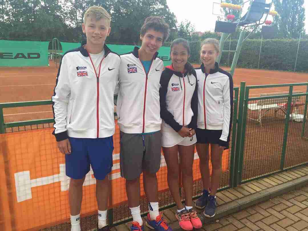 A young Emma Raducanu, wearing a white, blue, and red tennis jacket, stands with three other young tennis players on a court.