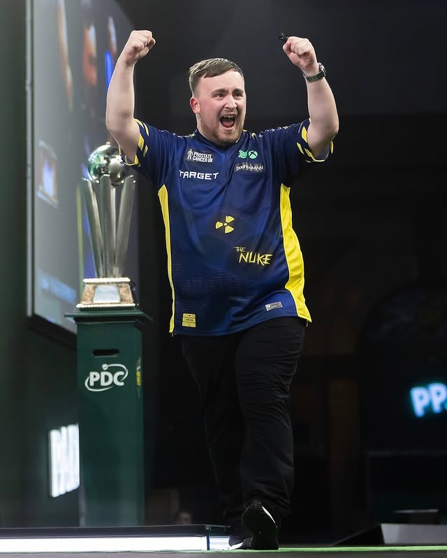 Luke Littler Celebrating with his arms raised, standing on a stage next to a trophy.