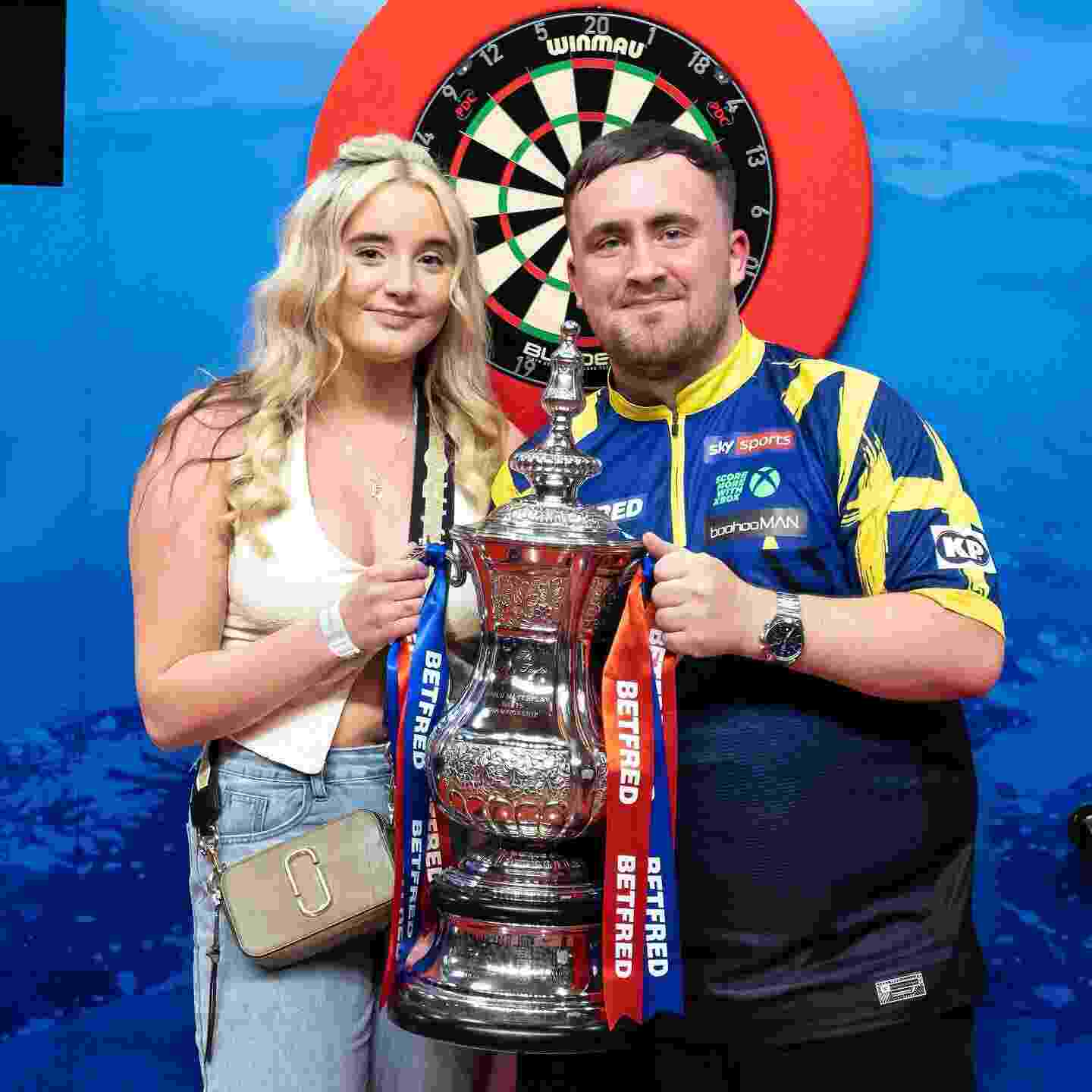 Luke Littler and his girlfriend, Faith Millar holding a trophy in front of a dartboard.