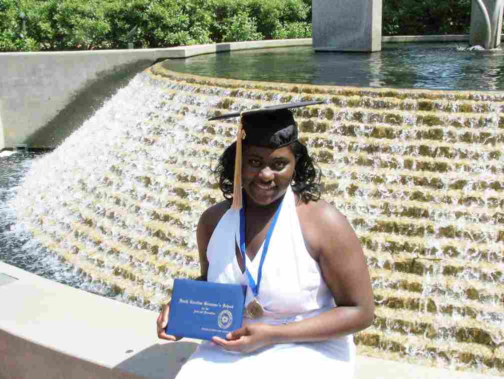 A young Danielle Brooks smiles, wearing a white dress, a graduation cap, and a medal, while holding her diploma in front of a water fountain.