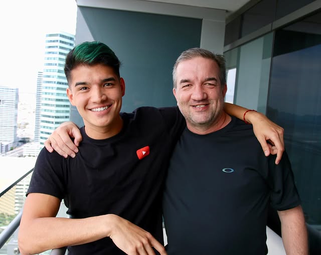 A smiling Alex Wassabi with his arm around his father, Adam Burriss standing on a balcony with a city view.