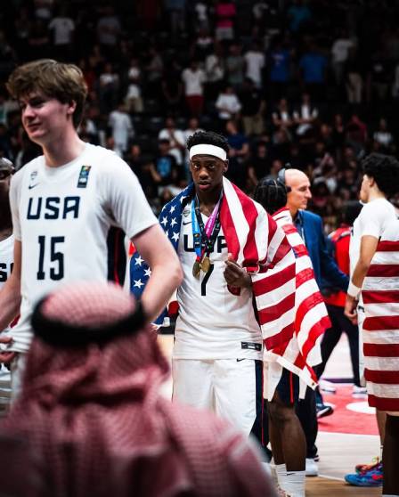 A.J. Dybantsa, wrapped in an American flag and wearing multiple gold medals, stands on the court with his teammates after a game.