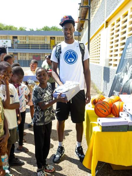 A.J. Dybantsa stands smiling and hands a shoebox to a young boy while surrounded by a group of children and a table of basketballs and shoe boxes.