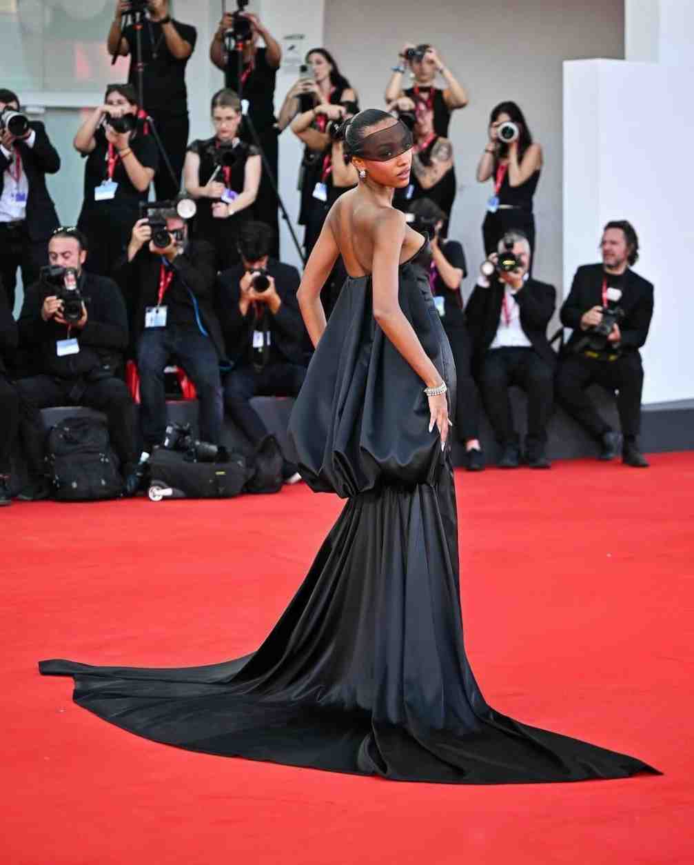 Sophie Wilde on the red carpet wearing a black gown with a long train and sheer veil, looking over her shoulder at photographers.