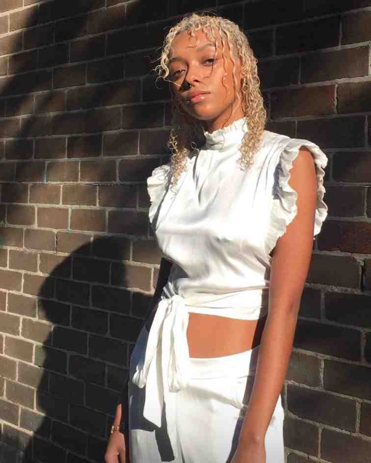 Sophie Wilde stands in front of a brick wall wearing a white, satin two-piece outfit with her blond hair in curls.
