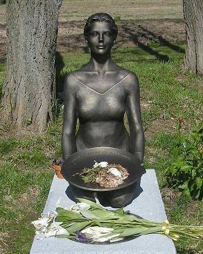 A bronze sculpture of a kneeling woman with a bowl on a headstone, surrounded by grass, trees, and flowers.