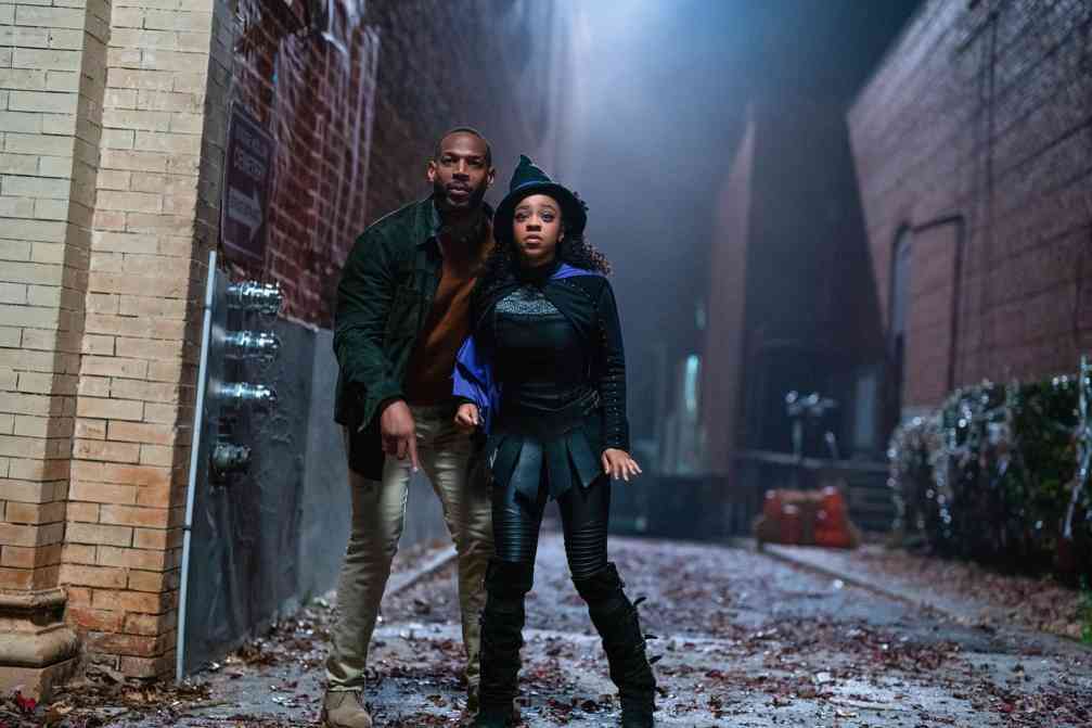 Priah Ferguson in a witch costume and Marlon Wayans in a casual jacket and pants stand together in a foggy alley, in a still from the film The Curse of Bridge Hollow.