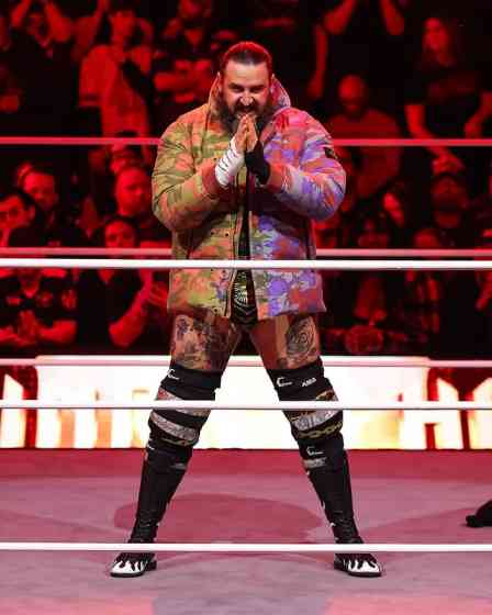 Professional wrestler Brody King, with a full beard and visible tattoos, stands in a wrestling ring, wearing a colorful camouflage jacket, wrestling trunks, and black boots.
