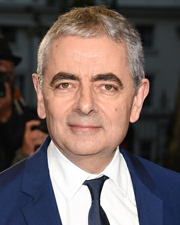 Rowan Atkinson, a middle-aged white man with short, greying hair, wearing a blue suit and a dark tie, looking directly at the camera with a neutral expression.