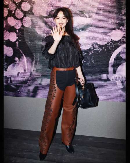 Lucie Zhang in a black top and brown chaps-style pants, holding a handbag and waving.