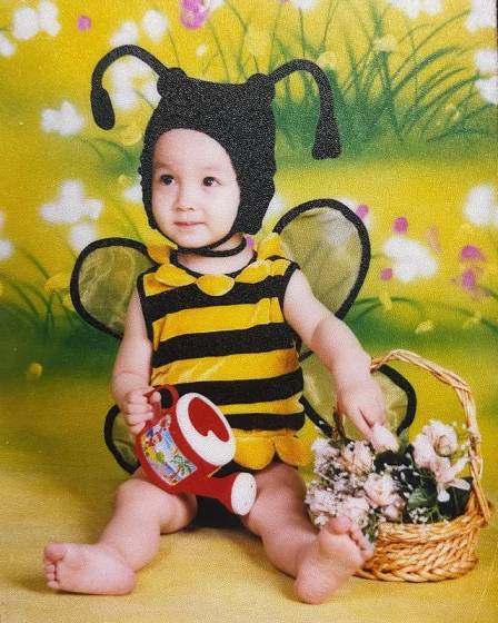 Young Lucie Zhang  dressed in a bee costume, holding a small watering can and a basket of flowers.