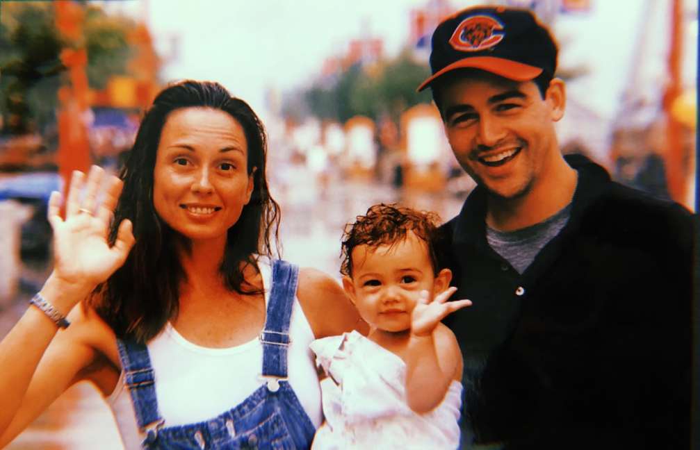 Kyle Chandler and Kathryn Macquarrie, holding a  young Sydney Chandler. Kathryn is wearing a denim overall and a white shirt while Kyle is wearing a baseball cap and al of them are waving at the camera.