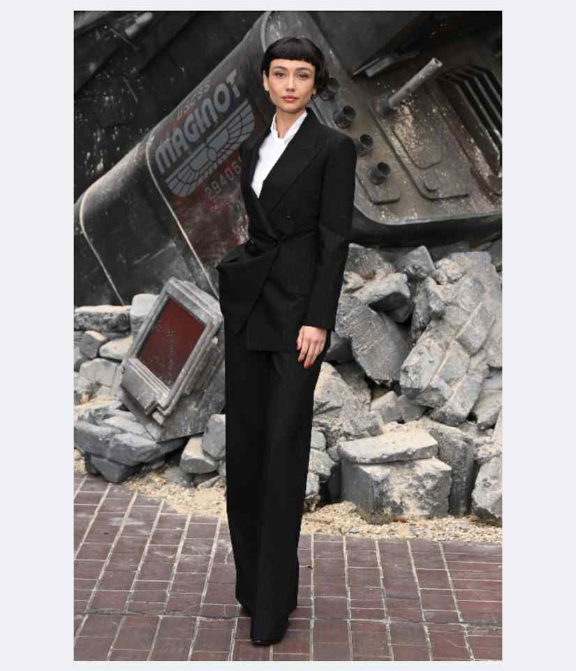 Sydney Chandler poses in a black pantsuit, with a white shirt and black heels, in front of a pile of rubble and a large, metallic structure.