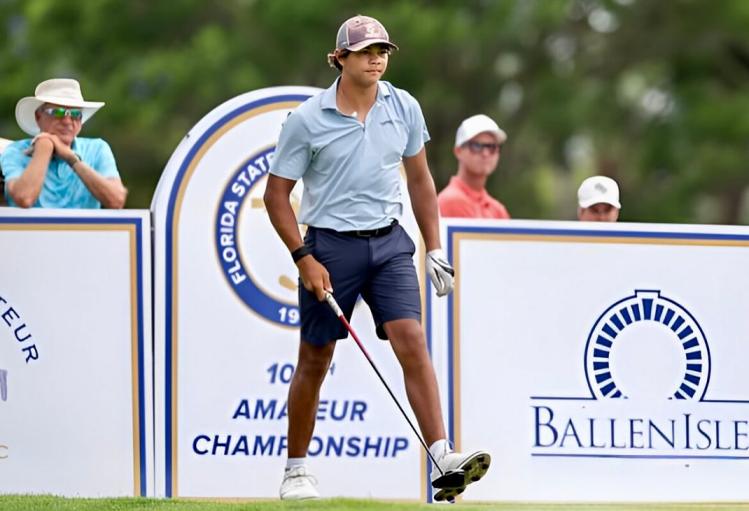Charlie Woods, dressed in a light blue polo and navy shorts, walks on a golf course with a club in his hand, a golf tournament sign visible in the background.