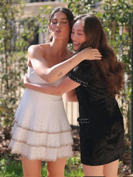 Havana Rose Liu and Gracie Abrams hug and smile outdoors, with Havana wearing a white dress and Gracie in a black one.