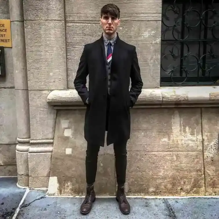 Actor Alex Breaux stands in front of a stone building, wearing a black coat, gray vest, and striped tie.