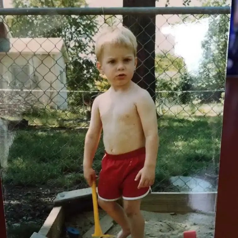 A young Alex Breaux as a toddler, standing in a sandbox with a toy shovel, wearing red shorts.