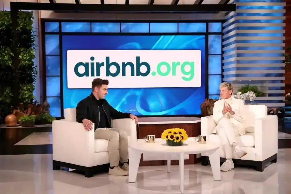 Brian Chesky, co-founder of Airbnb, sits on a talk show set across from Ellen DeGeneres with a large "airbnb.org" sign behind them.