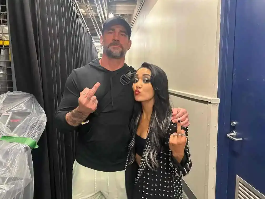 Roxanne Perez and  Cm Punk in a backstage hallway, both wearing black outfits and showing the finger.