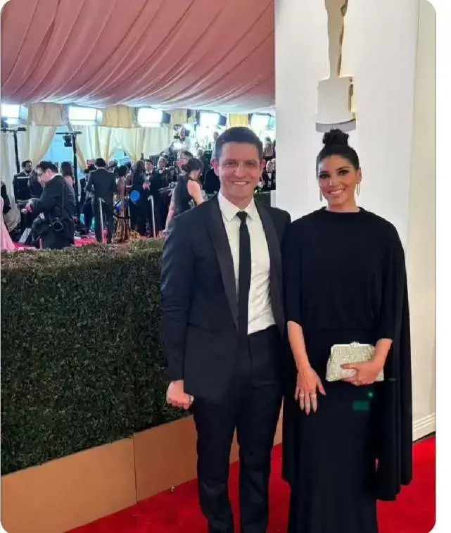 Amanda Setton and her husband, Ryan, are on the red carpet; Amanda, on the right, wears a black dress with a cape-like top and holds a silver clutch, while Ryan, on the left, sports a black tuxedo, and they are both smiling.