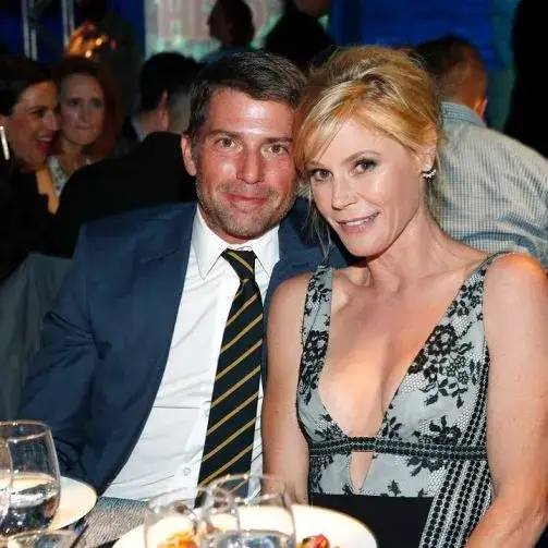 Julie Bowen and her ex-husband Scott Phillips smiling at an event, with Bowen wearing a V-neck, black and white patterned dress, and Phillips in a blue suit and striped tie.