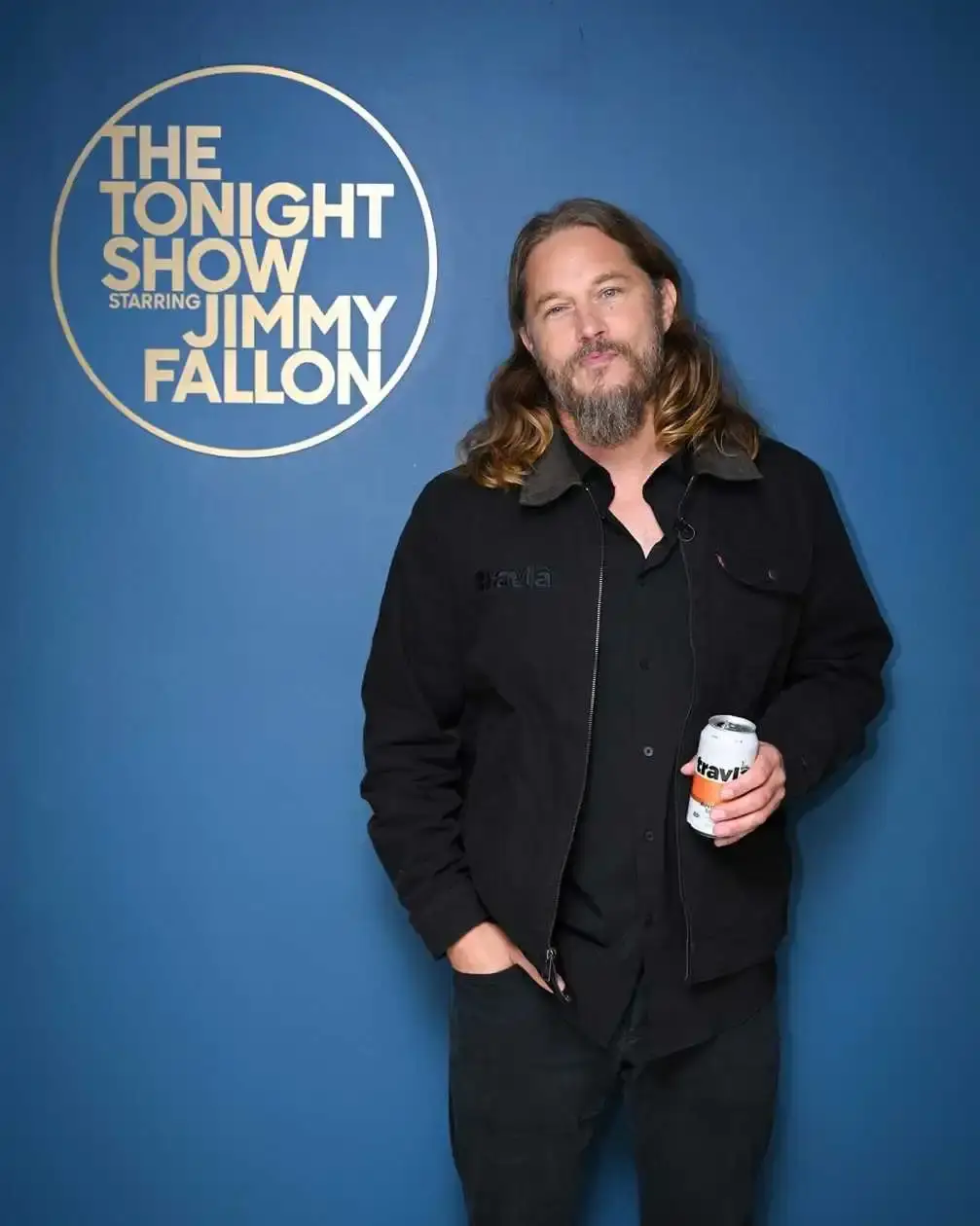 actor Travis Fimmel standing against a blue background with "THE TONIGHT SHOW STARRING JIMMY FALLON", has long hair and a beard and is holding a can in his hand.