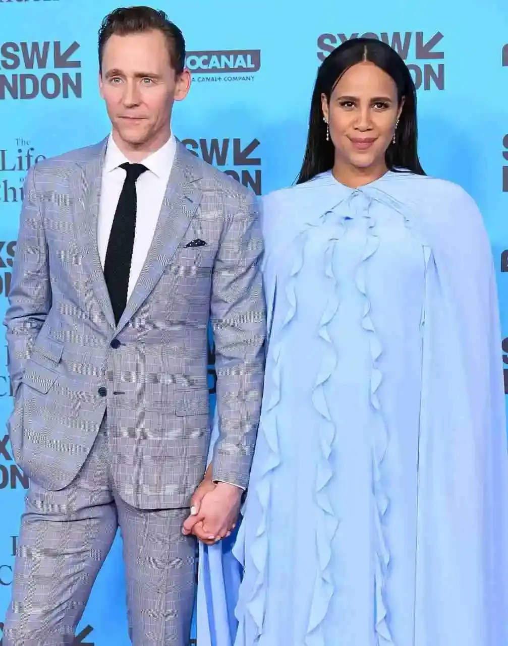 Zawe Ashton and her partner, Tom Hiddleston holding hands on a red carpet, with her in a light blue dress and him in a grey plaid suit.