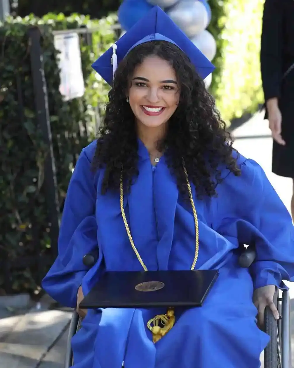 Marissa Bode smiles widely in a blue graduation cap and gown, holding her diploma in her lap while sitting in a wheelchair.