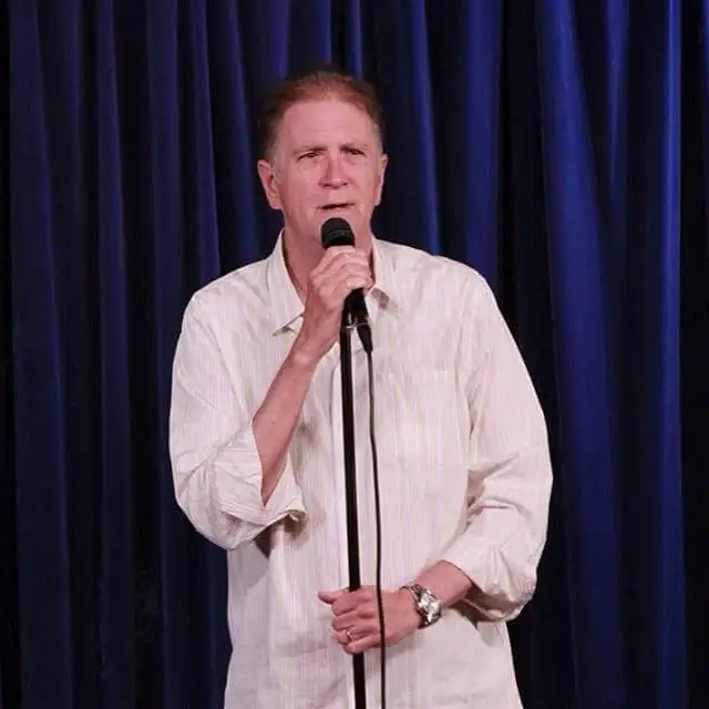 Allan Havey stands on a stage holding a microphone, wearing a light-colored striped shirt.