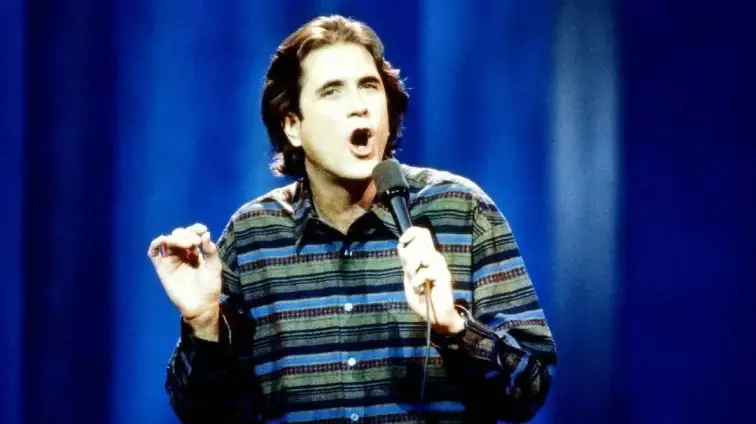 A young Allan Havey on stage holding a microphone, wearing a multi-colored striped shirt.