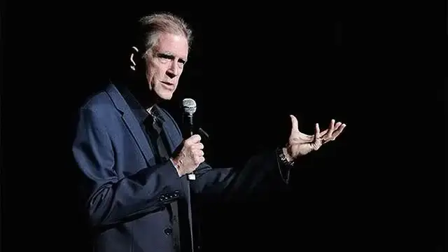 Comedian Allan Havey on a dimly lit stage, holding a microphone and gesturing with his free hand.