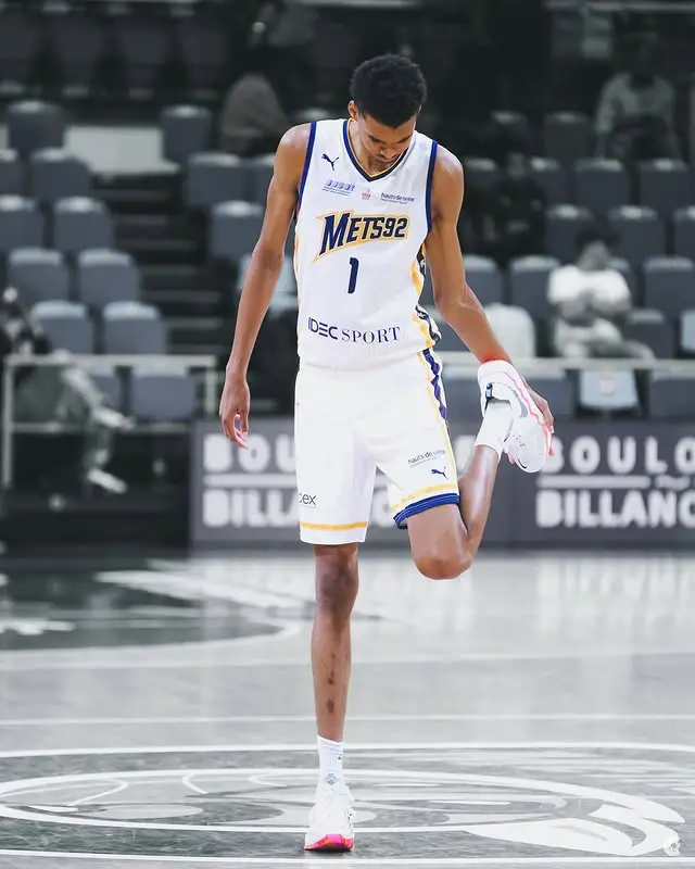 NBA player Victor Wembanyama is shown stretching his leg on a basketball court.