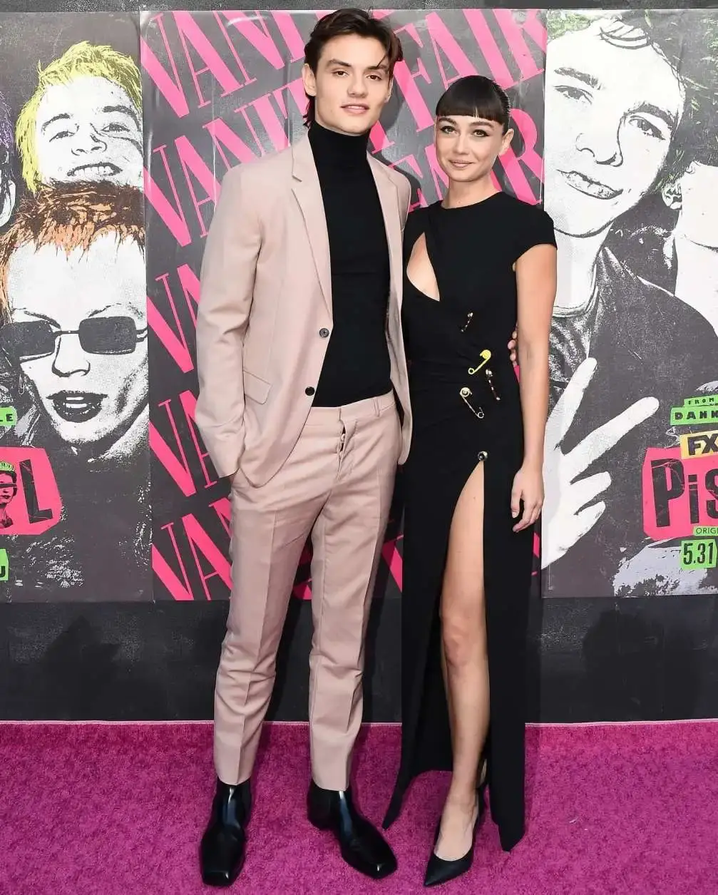 Sydney Chandler and Louis Partridge pose together on a pink carpet in front of a Pistol banner.