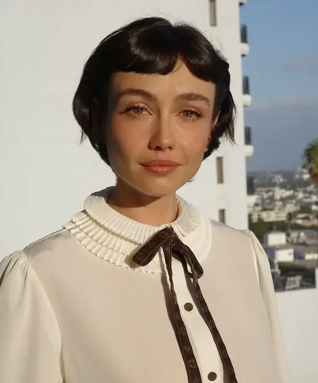Sydney Chandler smiles and looks at the camera while wearing a white blouse with a ruffled collar and a dark bow.
