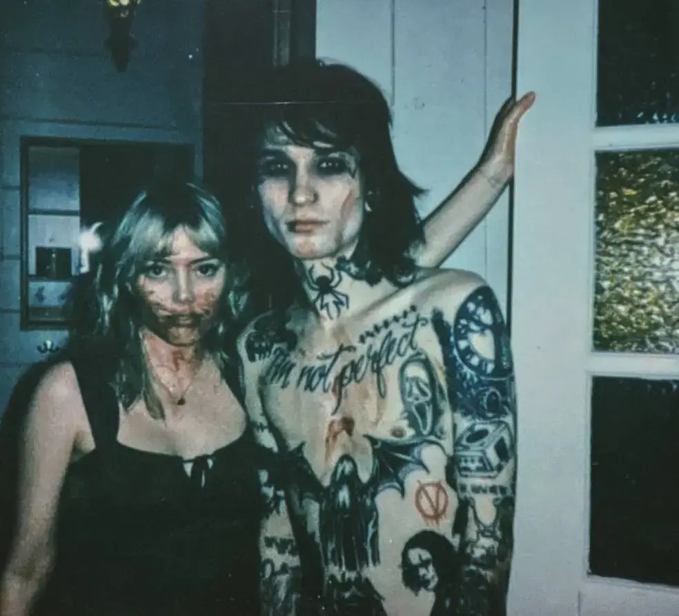 Grace Van Dien, with a bloody face and a black dress, stands next to Johnnie Guilbert, with face paint, a spider tattoo, and multiple other tattoos, who is holding a hand up against the wall.