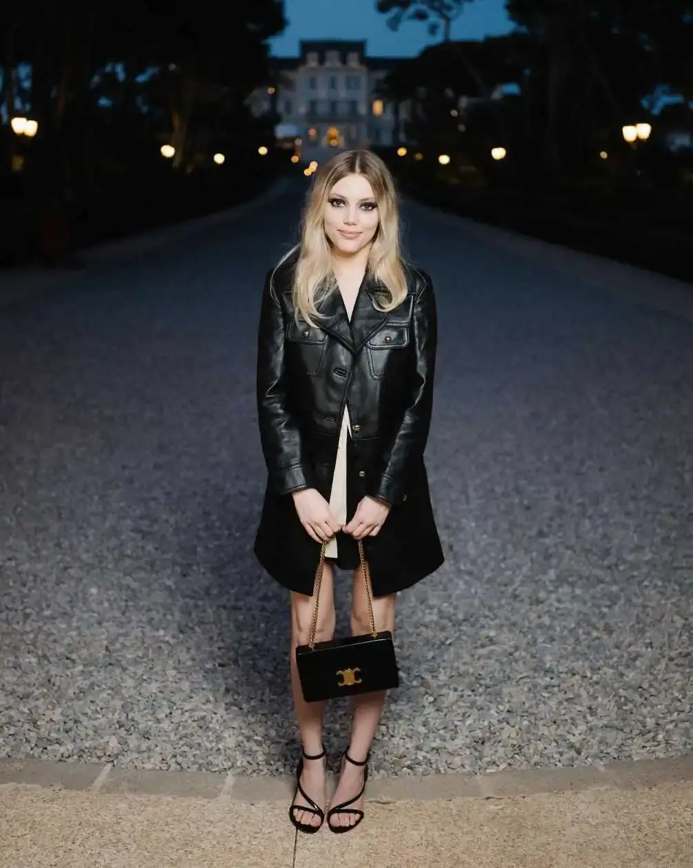 Grace Van Dien in a black leather jacket and a black dress holds a black clutch and smiles slightly, standing on a gravel path at dusk with a building in the background.