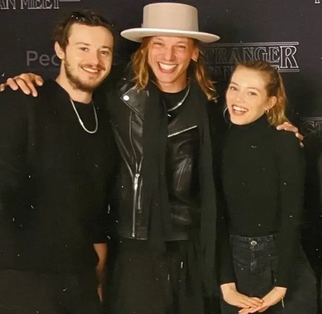 Joseph Quinn, on the left, with short brown hair and a silver chain, in the middle Jamie Campbell Bower has long blond hair and a top hat, while on the right Grace Van Dien has blonde hair in a ponytail.