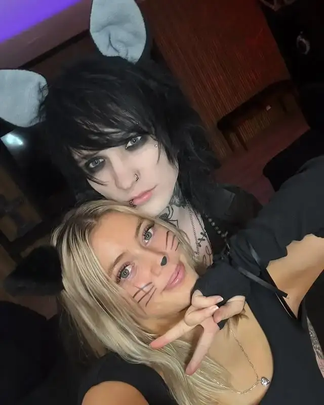 Grace Van Dien and Johnnie Guilbert, with black hair, both wearing cat ears, pose for a selfie. Likewise, Grace Van Dien has whiskers drawn on her face and is holding up a peace sign.