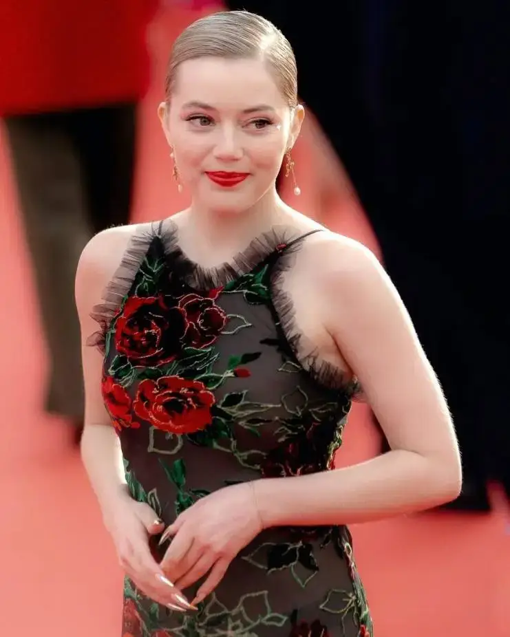 Grace Van Dien, with her hair pulled back and red lipstick on, wears a sheer black sleeveless dress with red flower embroidered on it.