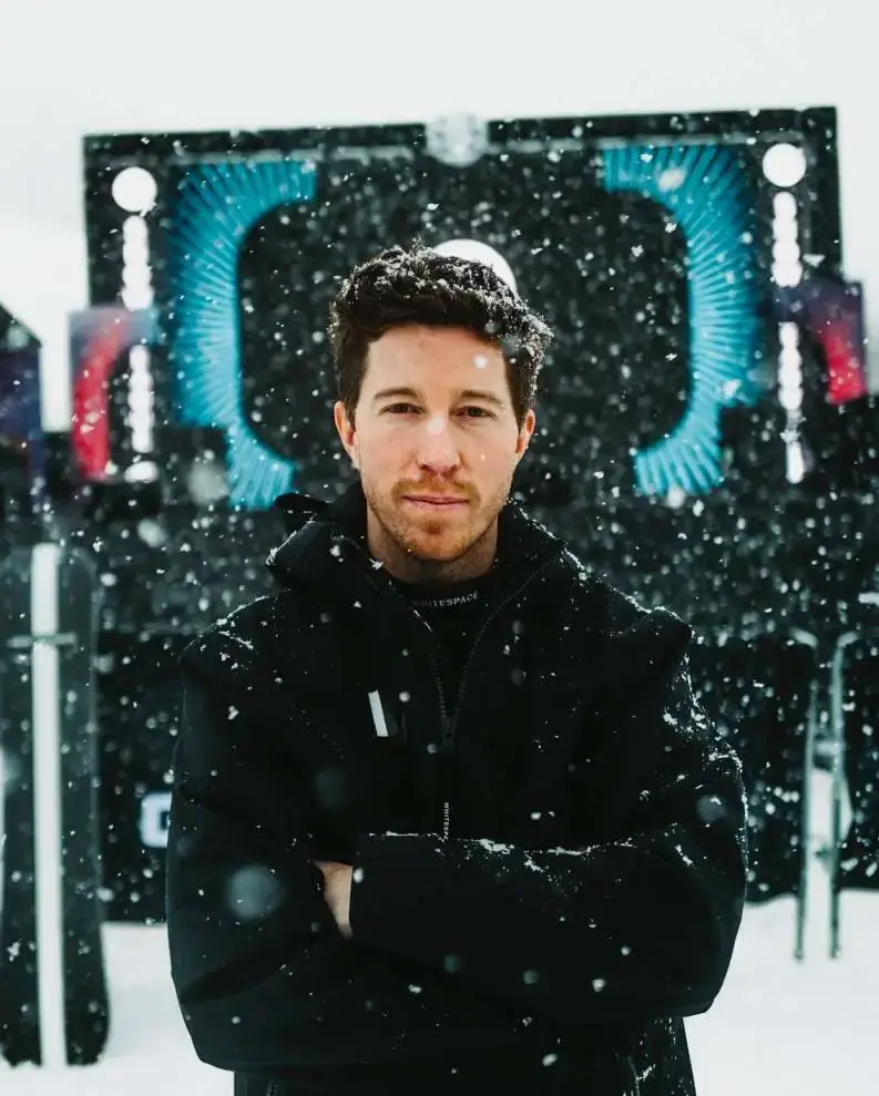 A picture of Shaun White, an Olympic snowboarder, wearing a black jacket with his arms crossed as snow falls around him.