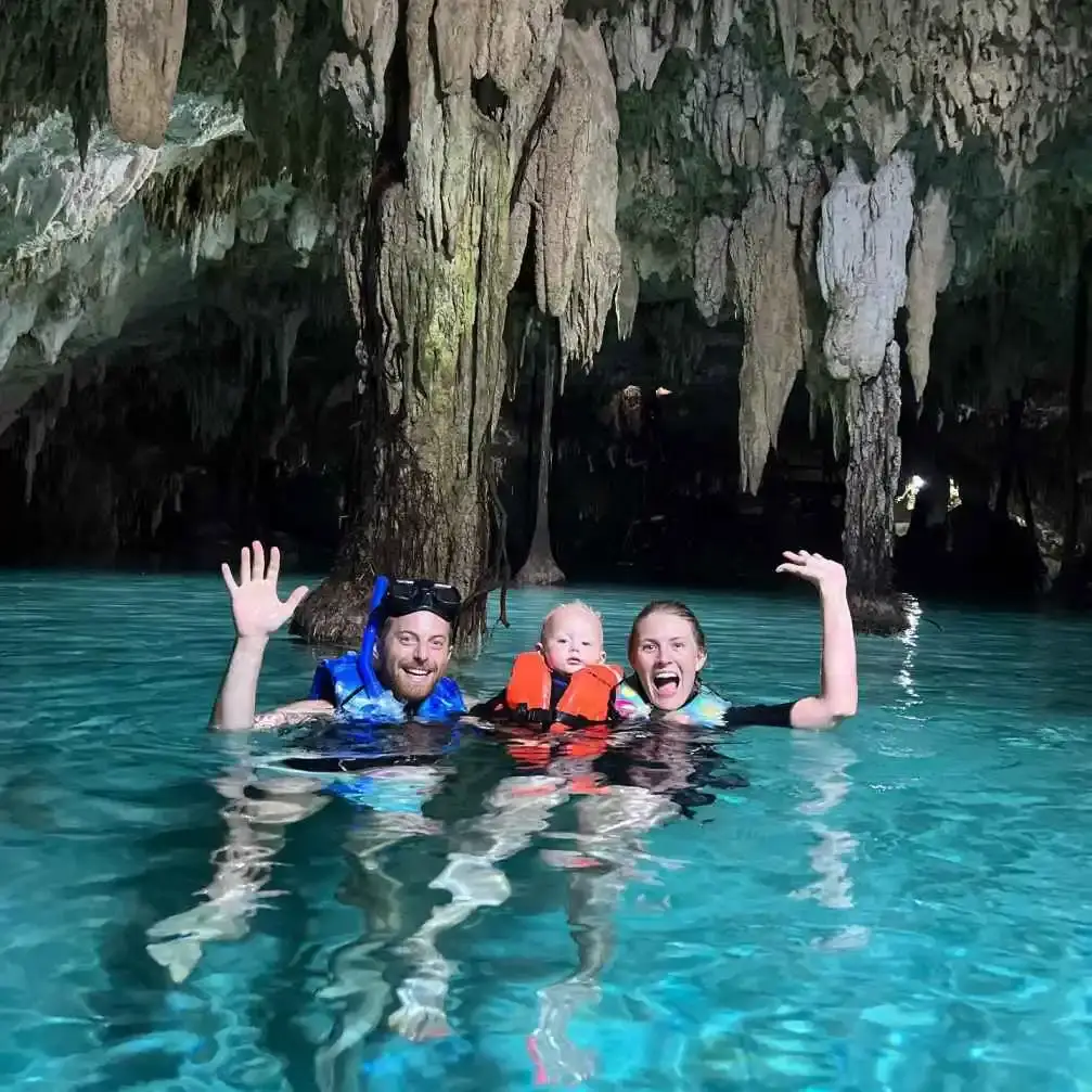 Ned Fulmer, his ex-wife Ariel Fulmer, and their child smiling and waving in a swimming at a cave, with stalactites hanging from the ceiling above them.