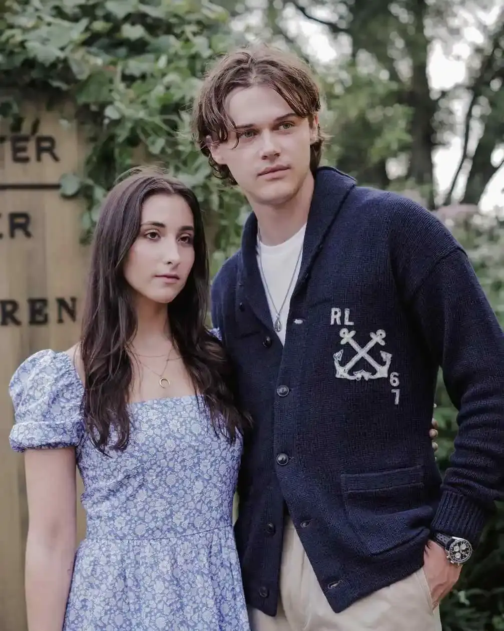 Christopher Briney and Isabel Machado stand close together, looking off to the side, with a wooden sign behind them.