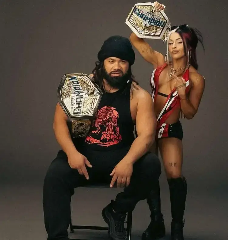 Jacob Fatu, with a beard and a black beanie, holds a championship belt, while a Zelina Vega wrestler in a red and black outfit stands behind him, holding another belt up.