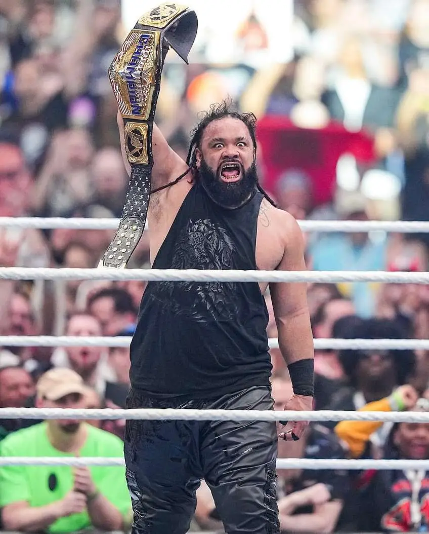 Jacob Fatu with a black tank top and pants stands in a wrestling ring, holding a championship belt above his head with a celebratory and intense expression.